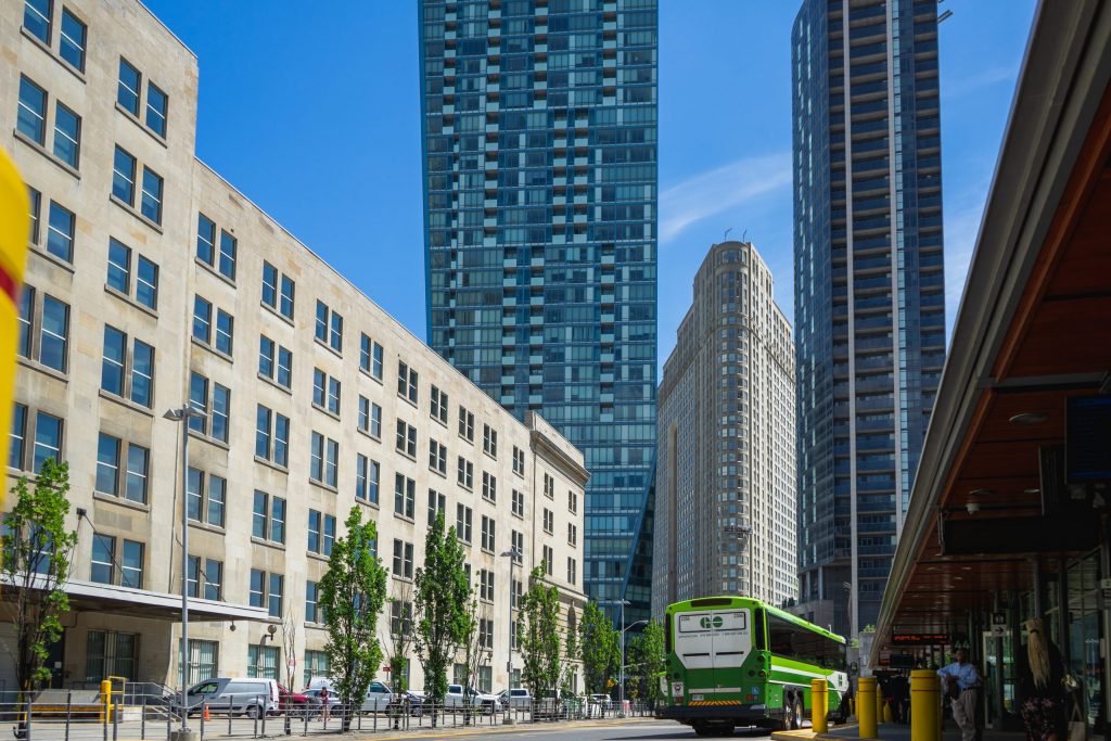 Two tall buildings in the background with a flat iron building in the middle. On the left is a midrise building, and on the right is a green and white GO bus.