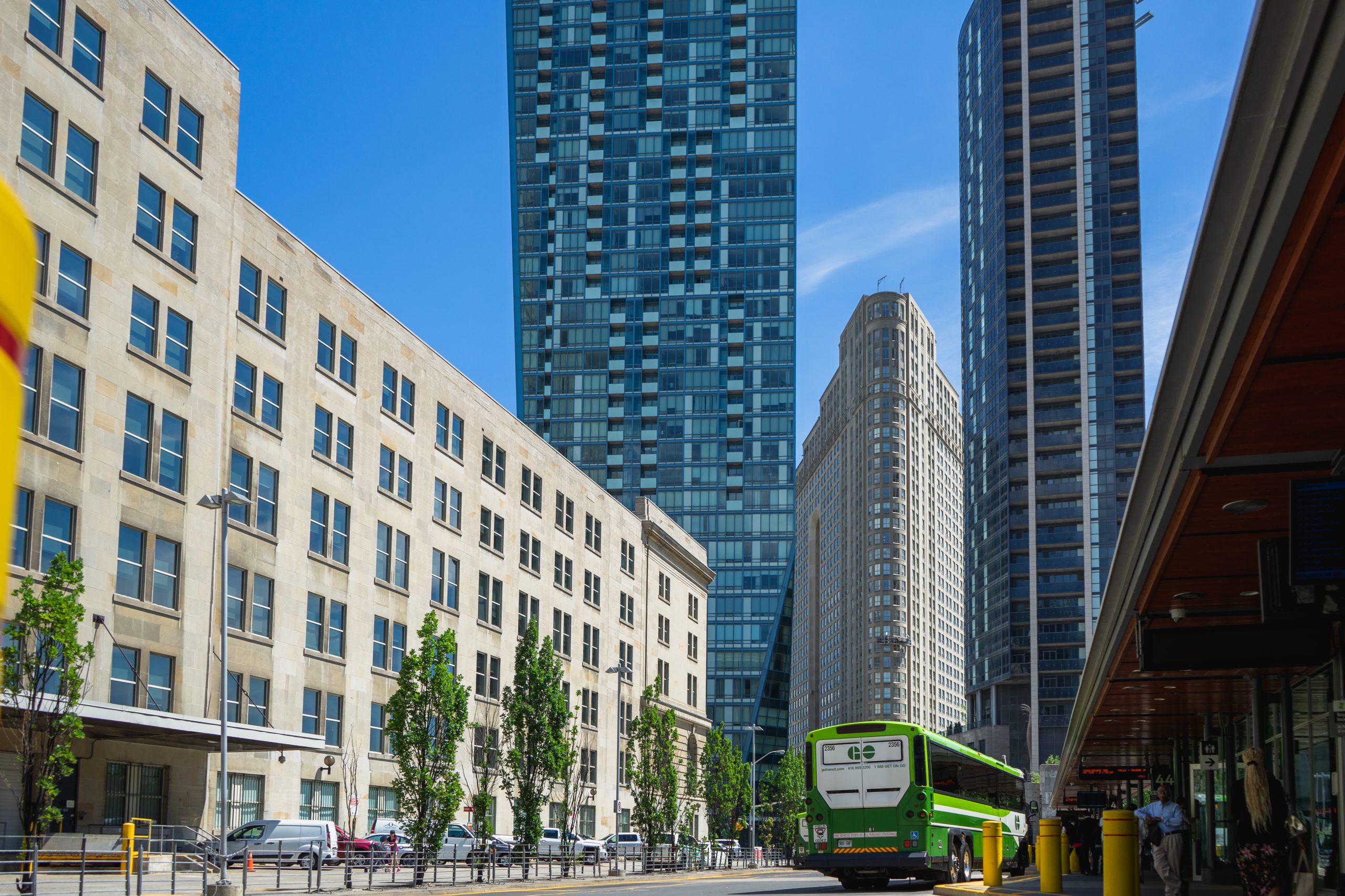 Two tall buildings in the background with a flat iron building in the middle. On the left is a midrise building, and on the right is a green and white GO bus.