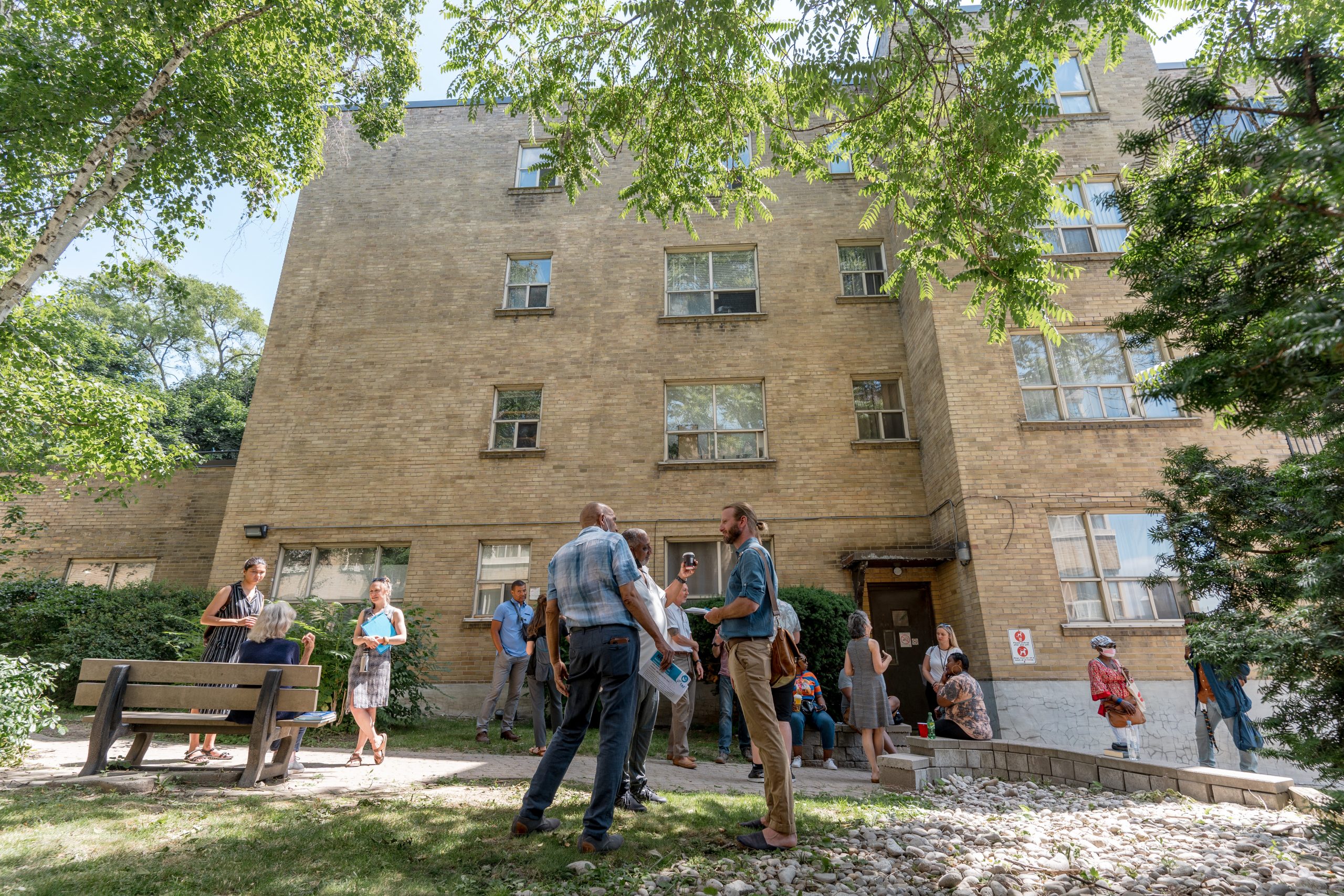 People are gathered in small groups in a green space in front of a multi-level residential building